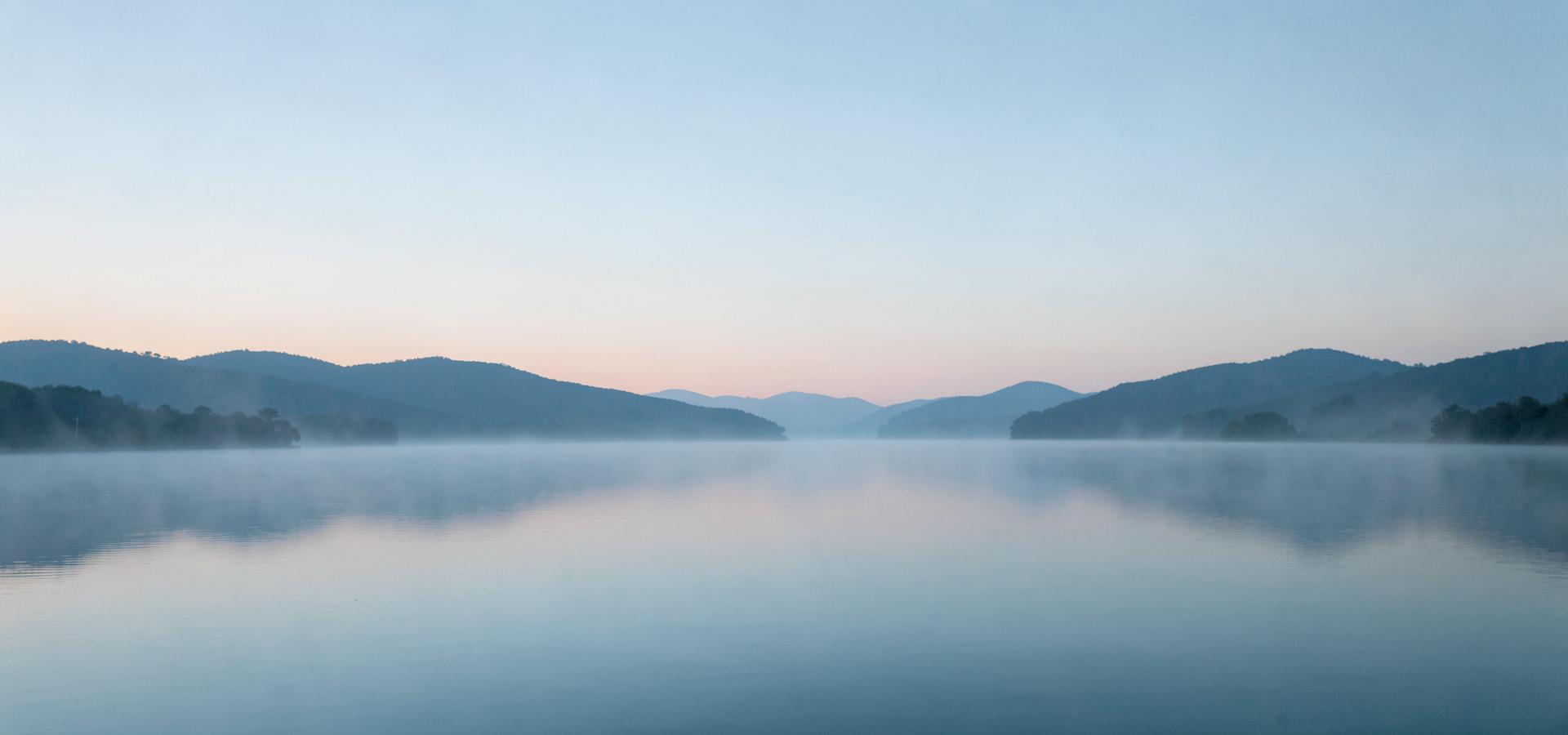 Lac calme au petit matin, brume sur l'eau et collines à l'horizon